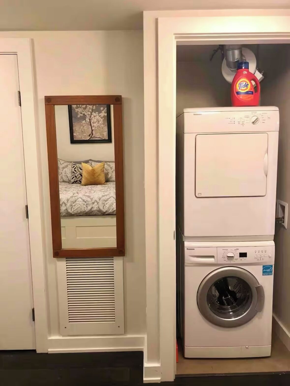 Ein Waschraum mit nebeneinander gestapelten Waschmaschine und Trockner, einem gerahmten Bild an der Wand und einem Bett mit gemusterten Betttextilien.
