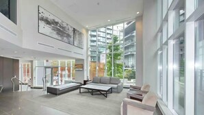 A spacious lobby with a modern design, featuring a seating area with sofas and a coffee table, large windows offering views of the surrounding buildings, and a reception desk. - Beautiful Downtown Condo- A/C, Parking, Laundry, Great Location! (Vancouver)