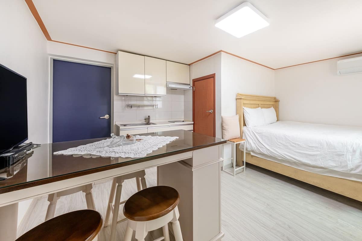 A compact hotel room with a kitchenette, a bed, a TV, and a glass dining table.