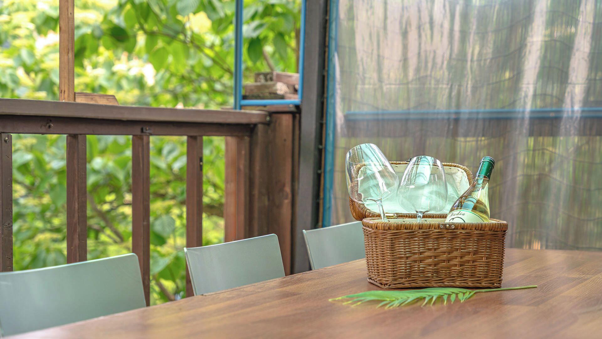 A wooden table with a wicker basket containing wine glasses and a bottle, set against a backdrop of greenery.