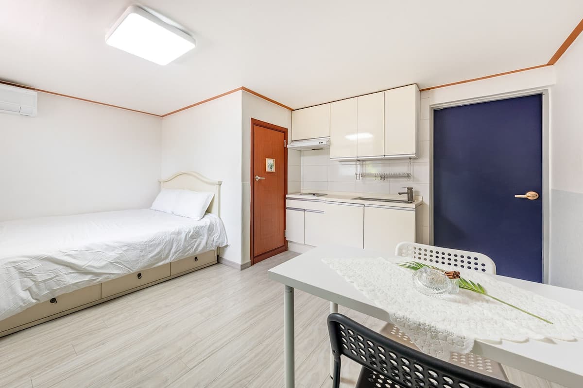 A compact room with a bed, a small kitchenette, a desk with a chair, and a door.