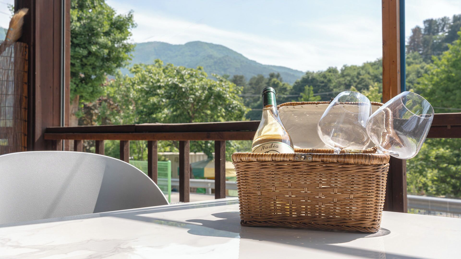 Una cesta de mimbre sobre una mesa de balcón con una botella y copas, con vistas a un paisaje montañoso.