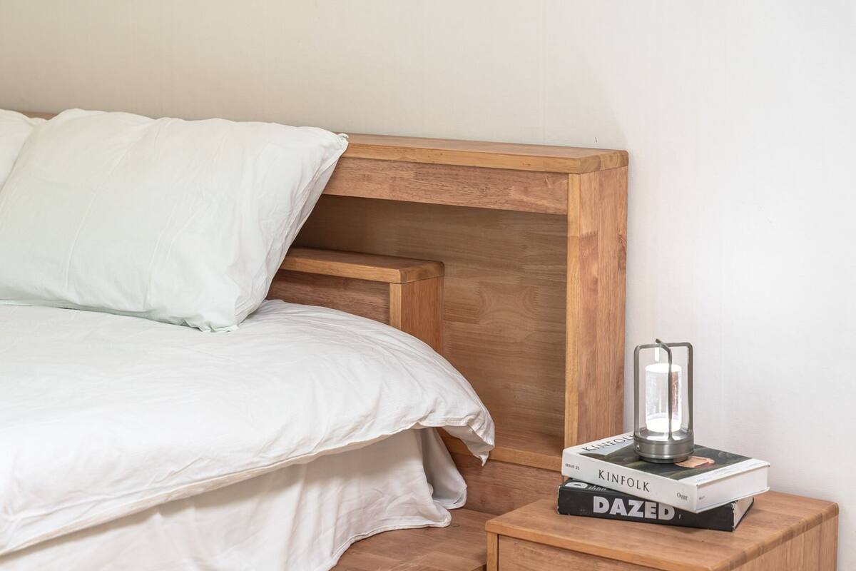 A bed with white bedding, a wooden headboard, and a bedside table with books and a lamp.