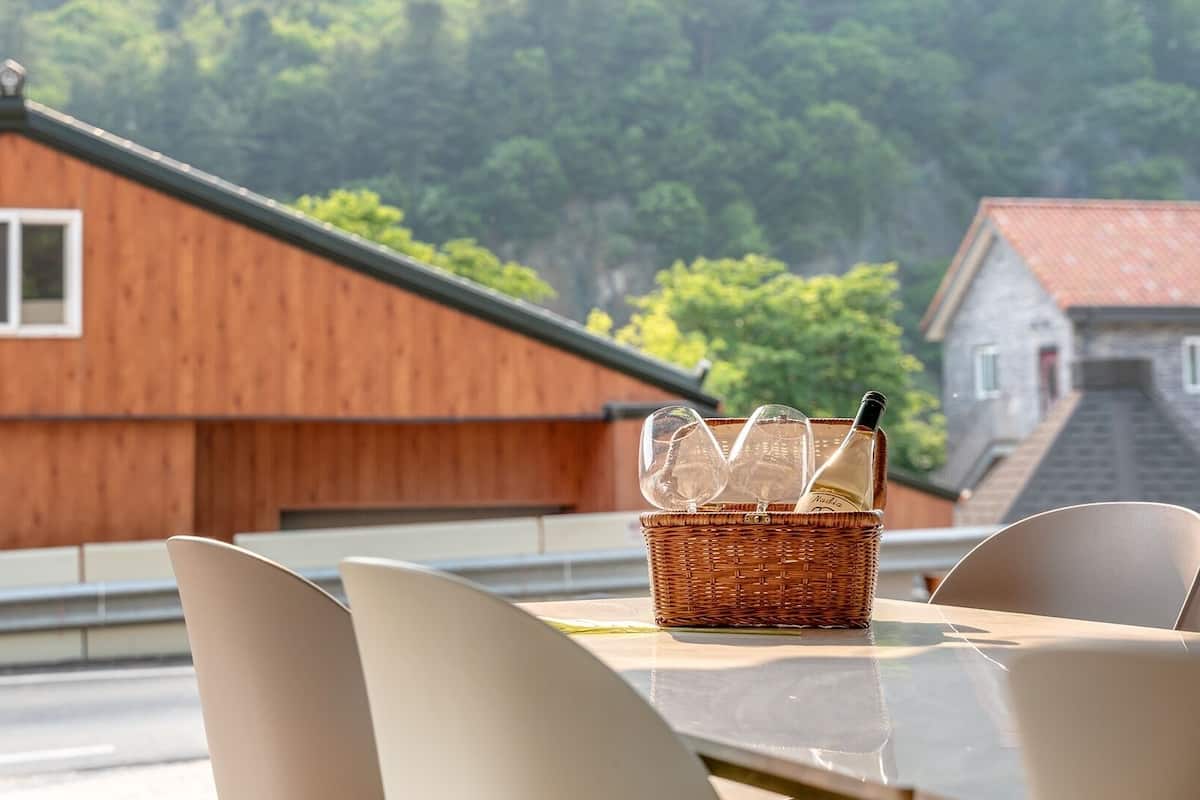 A table with a wicker basket containing wine glasses and a bottle, set against a backdrop of a wooden building and a stone wall.