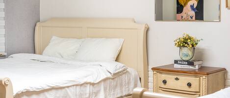 Una cama bien hecha con ropa blanca, una mesita de noche con un jarrón de flores y un cuadro en la pared.