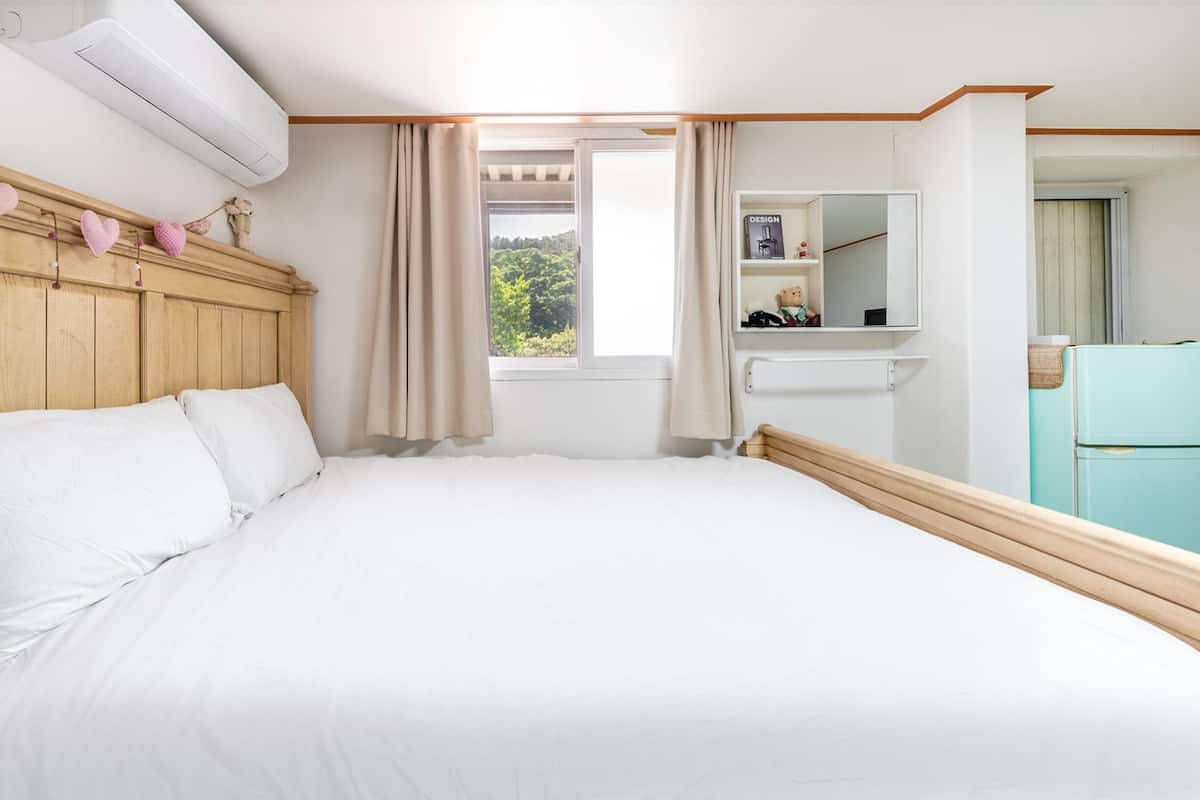 A bedroom with a wooden headboard, a bed with white linens, a window with curtains, a mirror, and a teal-colored refrigerator.