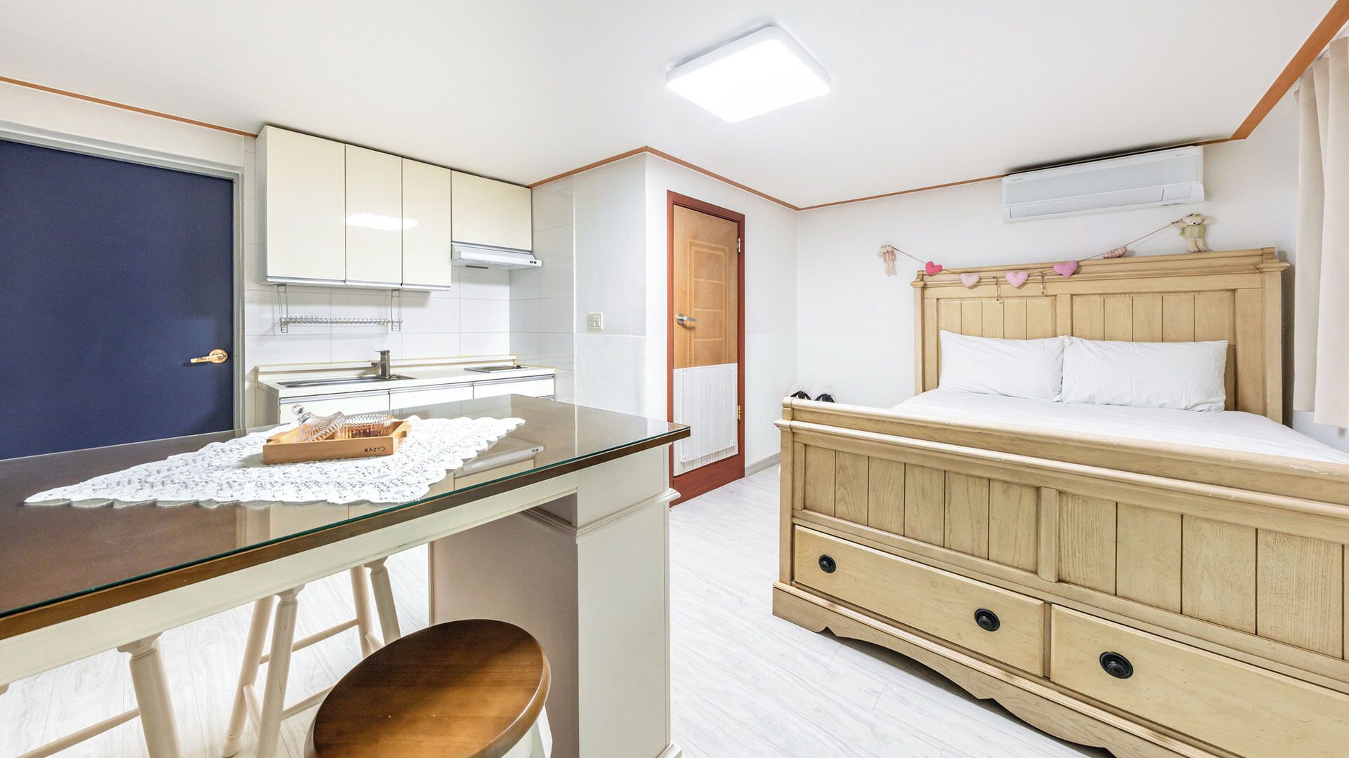 A compact room with a kitchenette, a bed with a wooden headboard, and a small table with a chair.