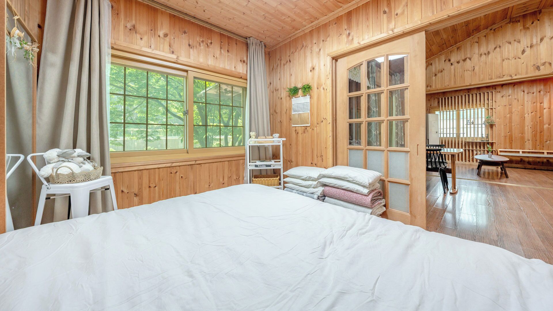 A wooden room with a bed, a chair, a table, and a window with a view of trees.