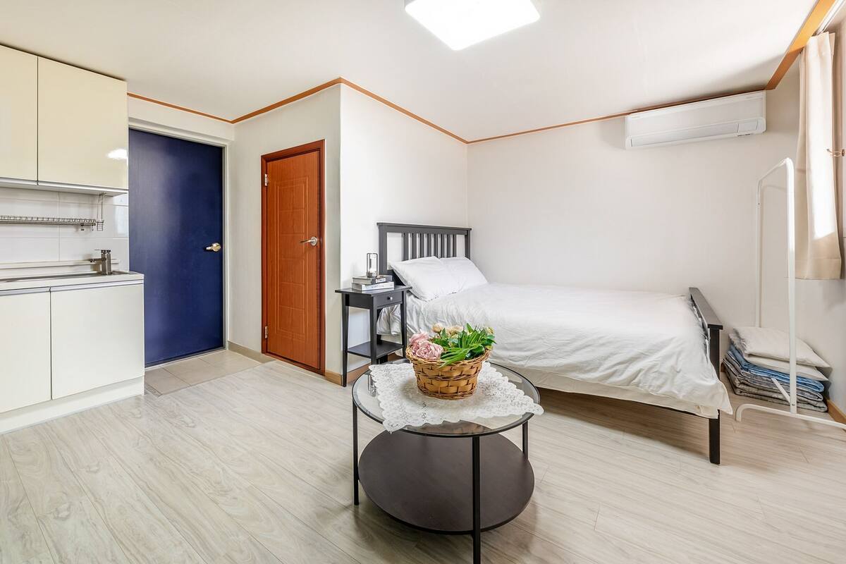 A compact room with a bed, a small table with a basket of flowers, a bedside table with a book, a blue door, and a kitchenette.