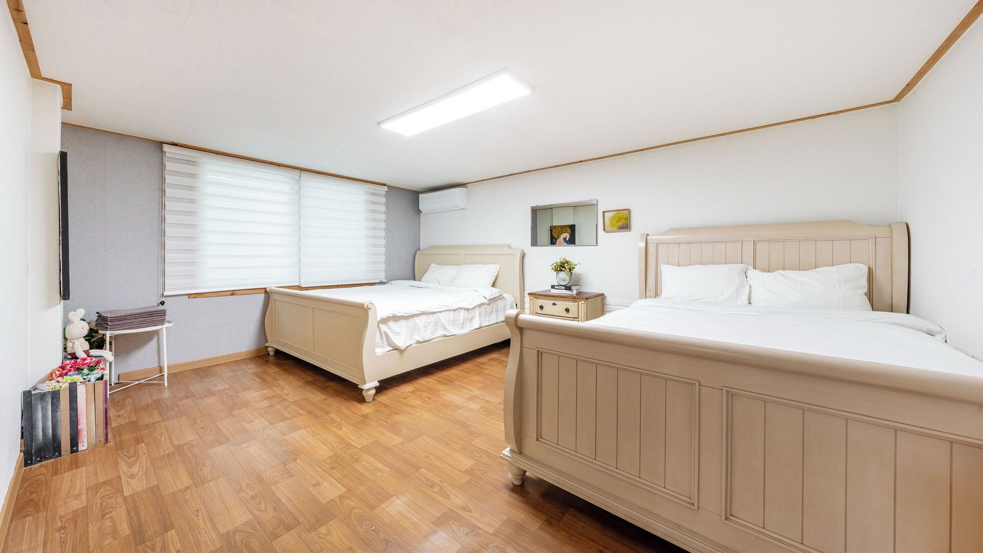 A hotel room with two beds, a bedside table with a lamp, a window with blinds, and a radiator.