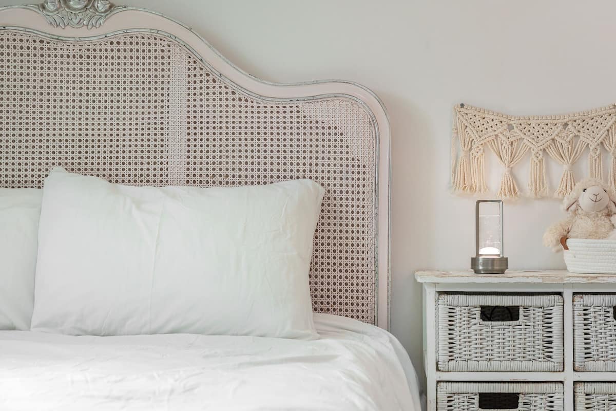 A bed with white bedding, a woven headboard, a macrame wall hanging, a lamp, and wicker baskets with stuffed animals.