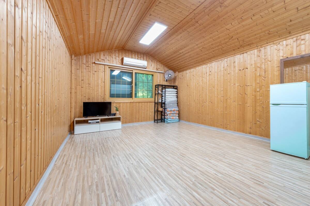 A spacious room with wooden walls and ceiling, a flat-screen TV on a white cabinet, a white refrigerator, and a window with blinds.