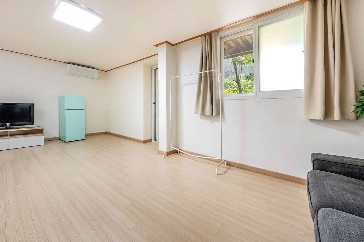 A living room with a flat-screen TV, a light blue refrigerator, a grey sofa, and a window with curtains.