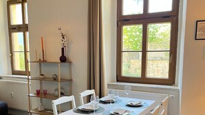 Un comedor con una mesa blanca dispuesta para cuatro personas, rodeada de sillas blancas con cojines oscuros. Una estantería de madera con adornos está contra la pared, y se ve una ventana con vistas a zonas verdes.
