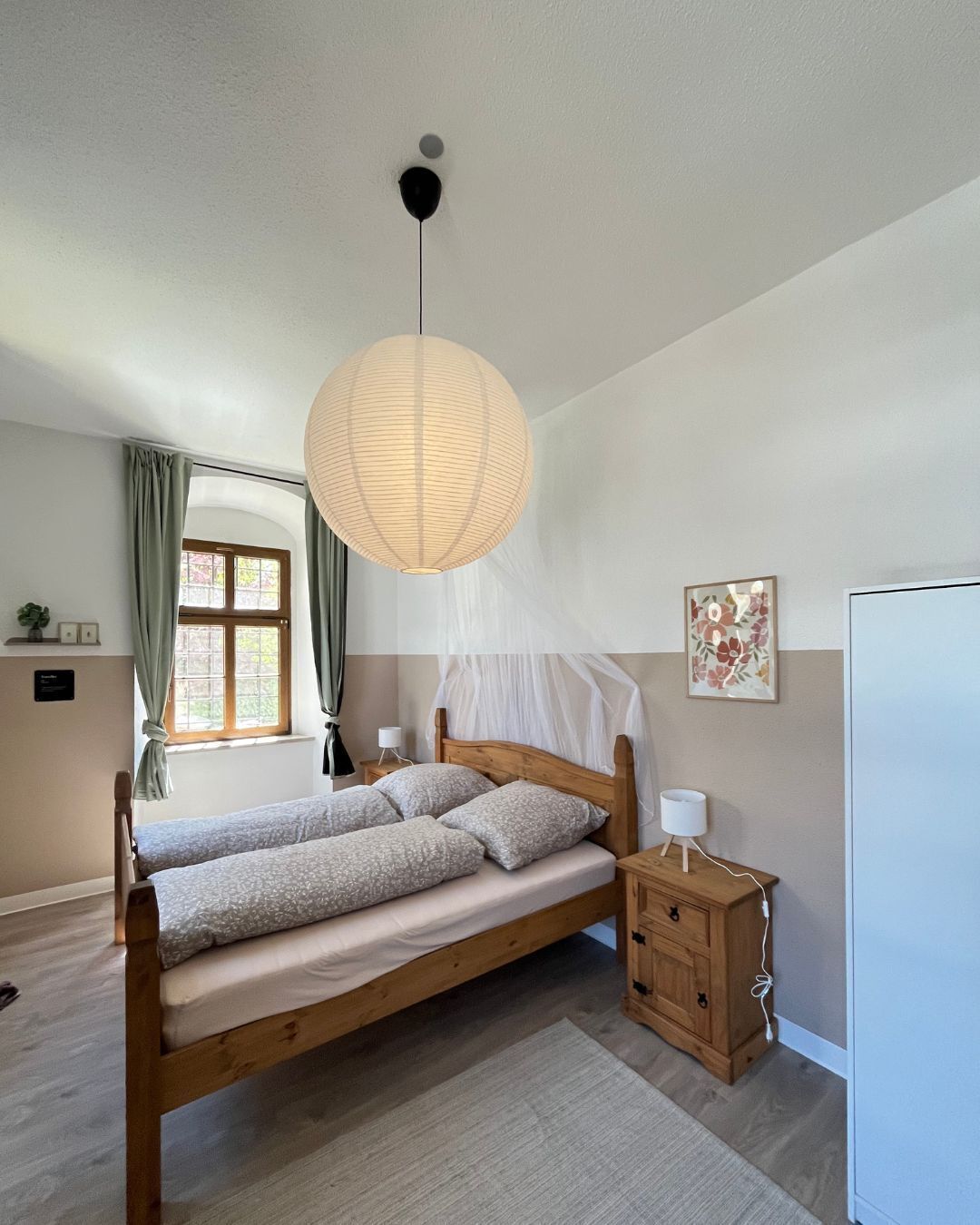 A bedroom with a wooden bed, a bedside table with a lamp, a hanging paper lantern, a window with curtains, and a wall-mounted picture.