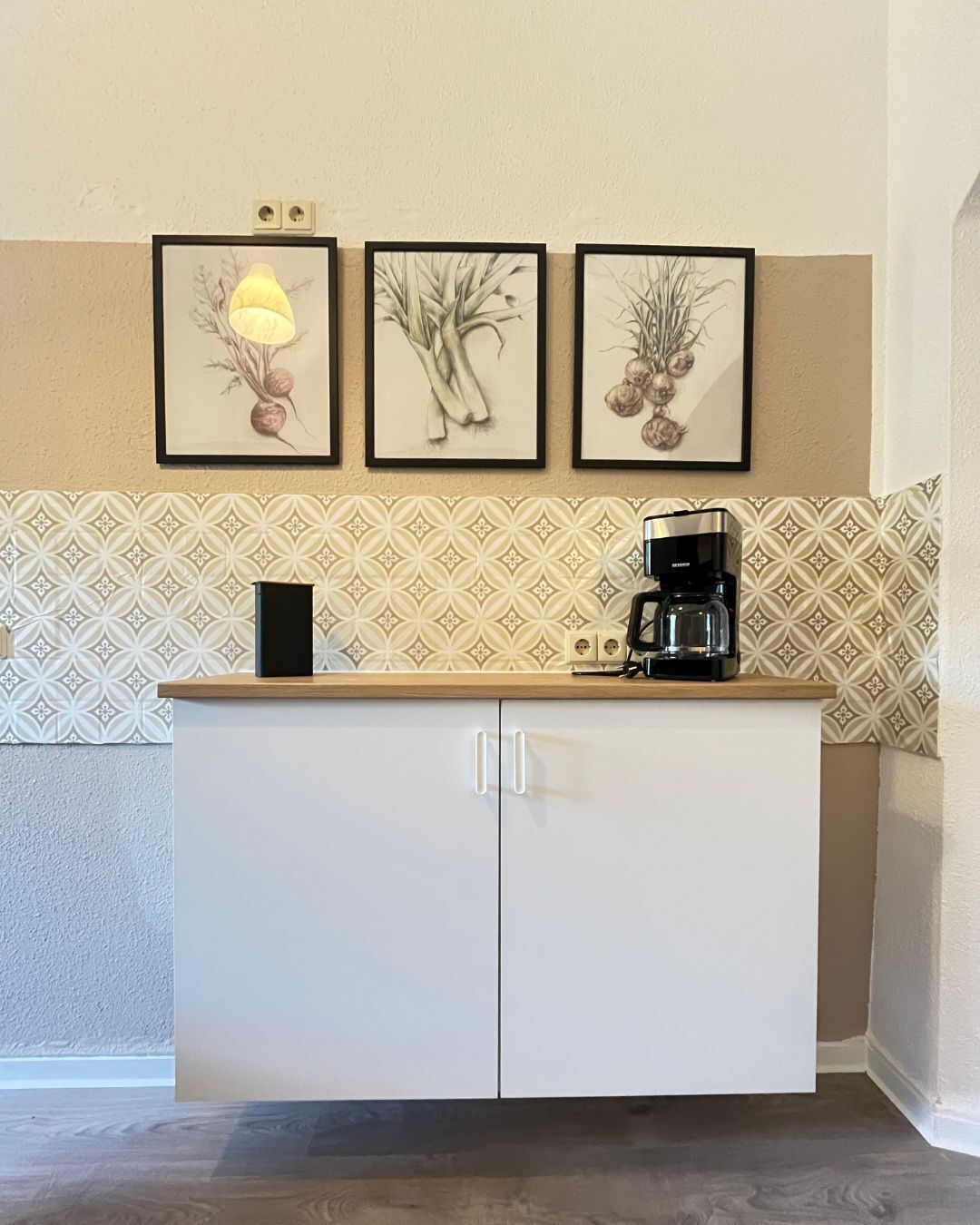 A kitchen with a white cabinet, a coffee maker, and three framed botanical prints on the wall.