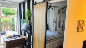 A room with a bed, a desk with a laptop, a plant, and a sliding door leading to a balcony.