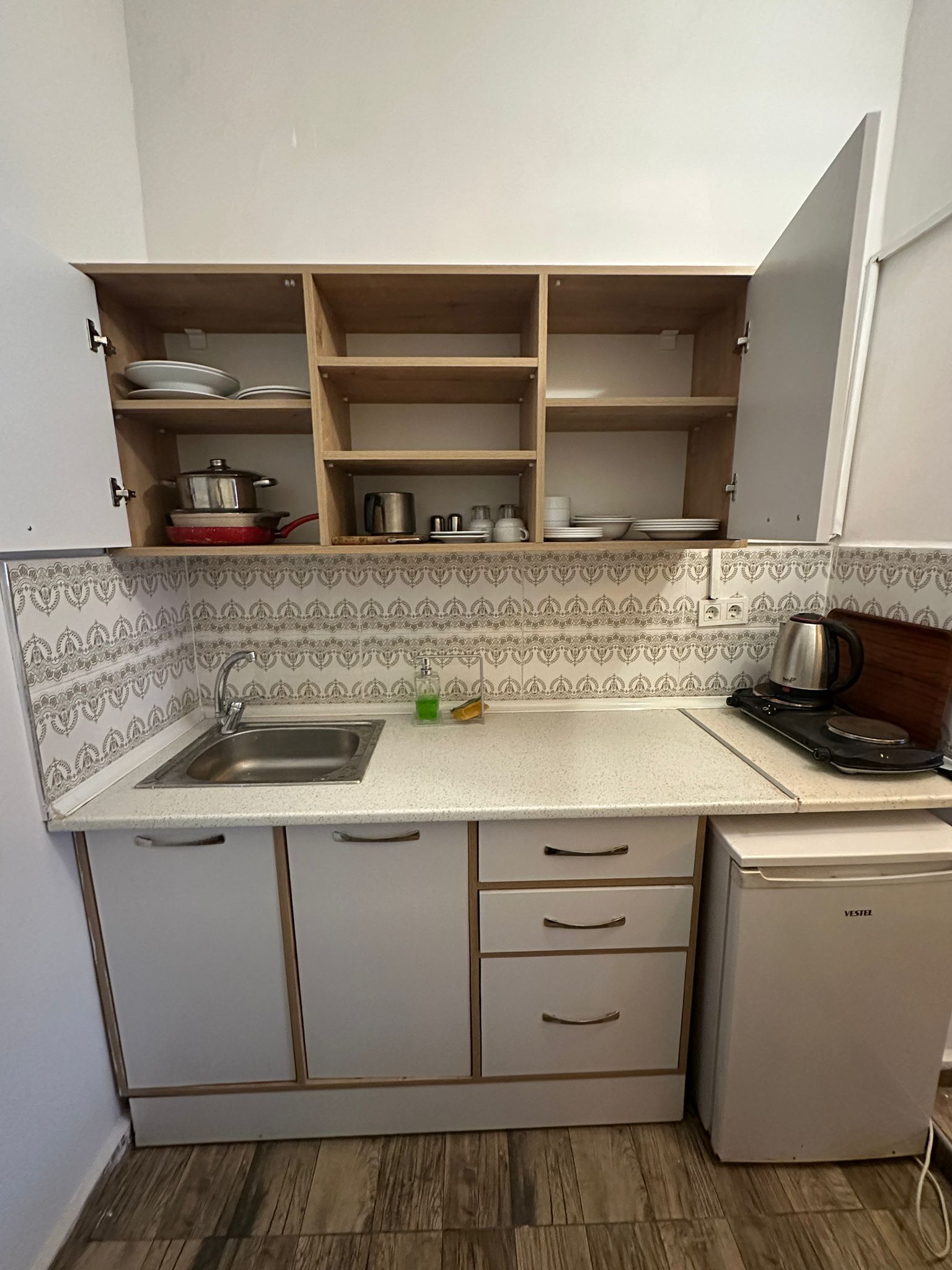 Een compacte keuken met een spoelbak, een kleine koelkast en houten planken boven het aanrecht.