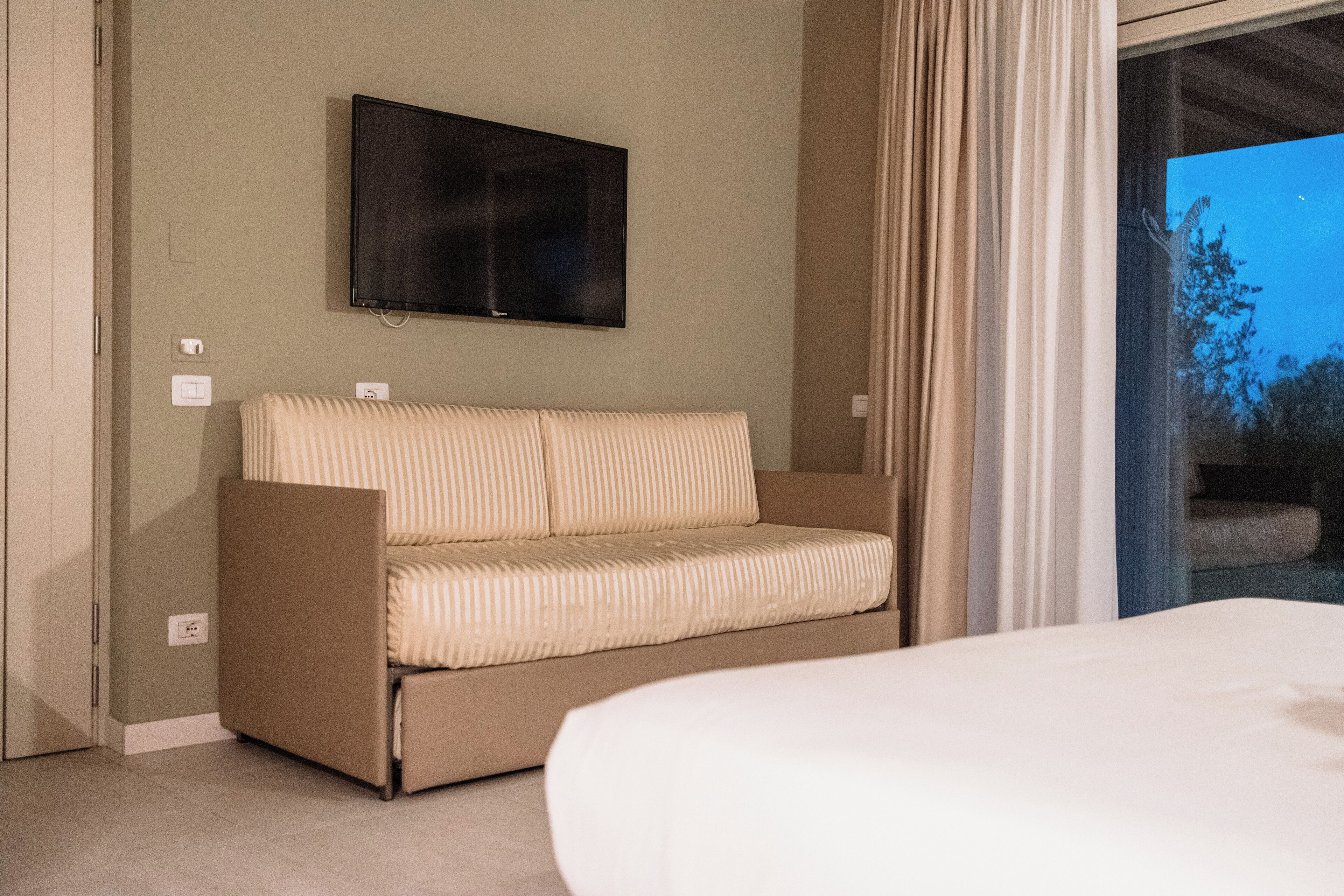 A hotel room with a sofa, a bed, a flat-screen TV mounted on the wall, and a view of the outdoors through a large window.