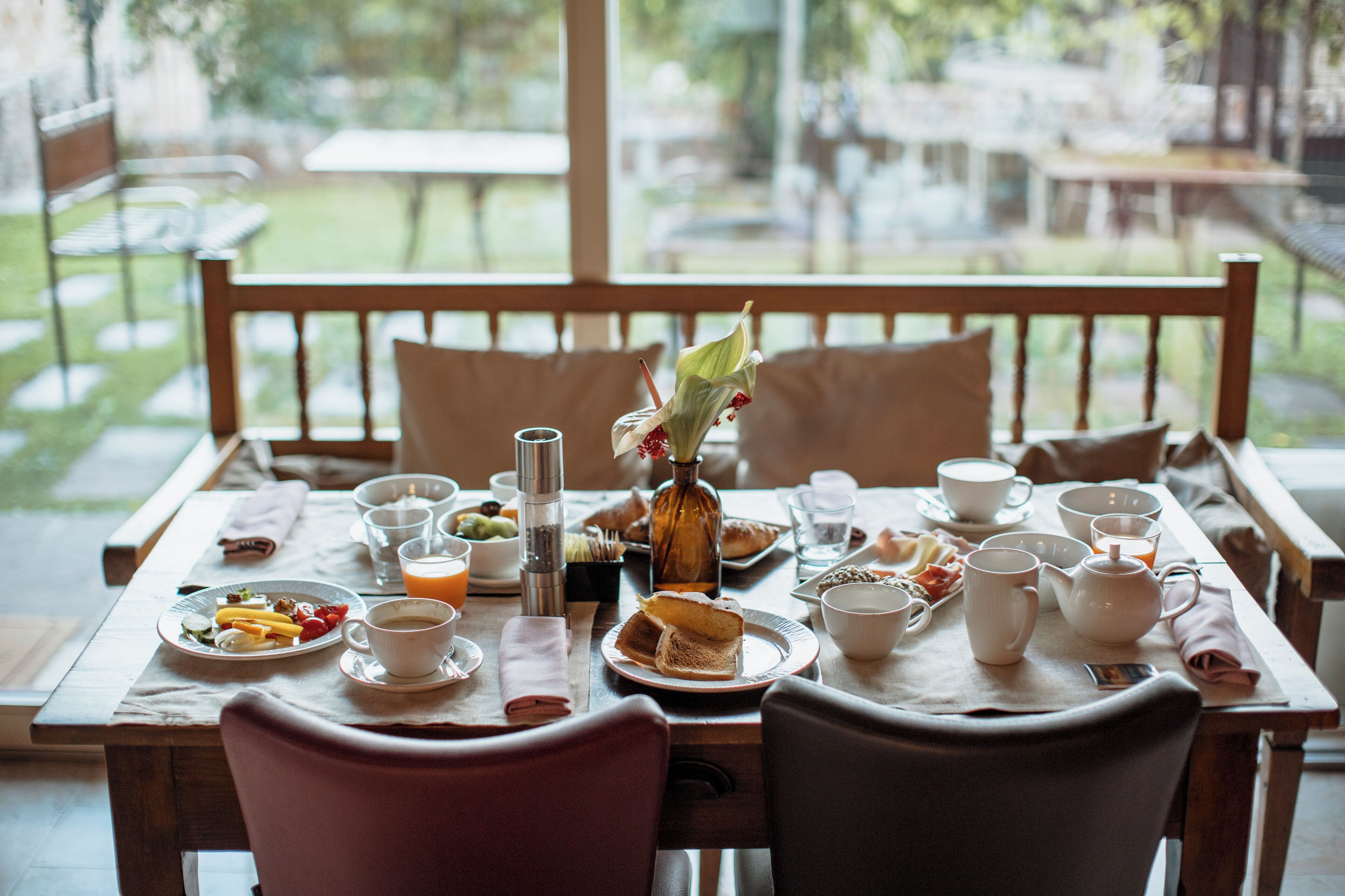 A breakfast table set with plates, cups, and a teapot, overlooking a garden.