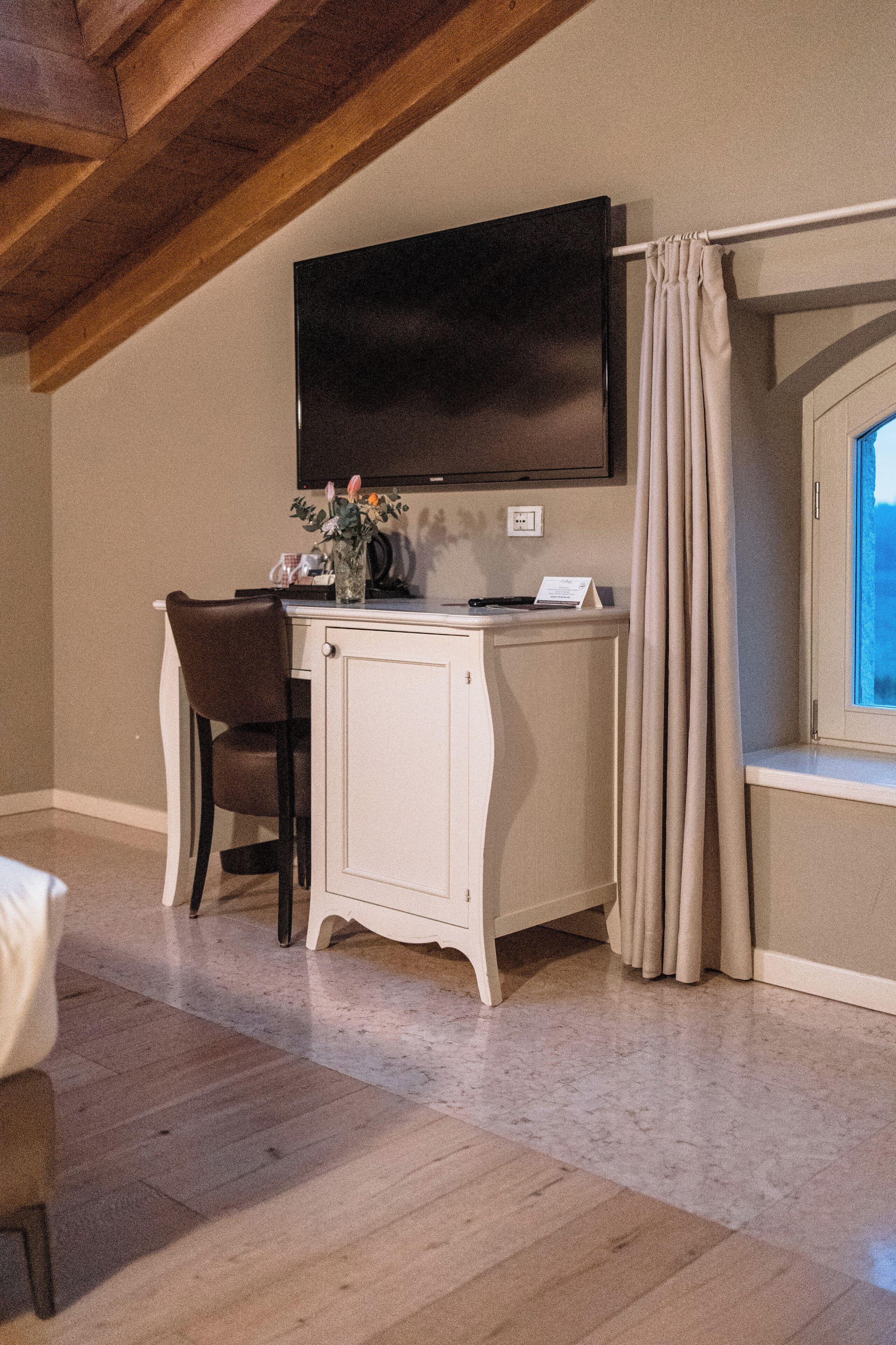 Une pièce avec un bureau blanc, une chaise marron, un téléviseur à écran plat fixé au mur, une fenêtre avec des rideaux et une poutre en bois au plafond.