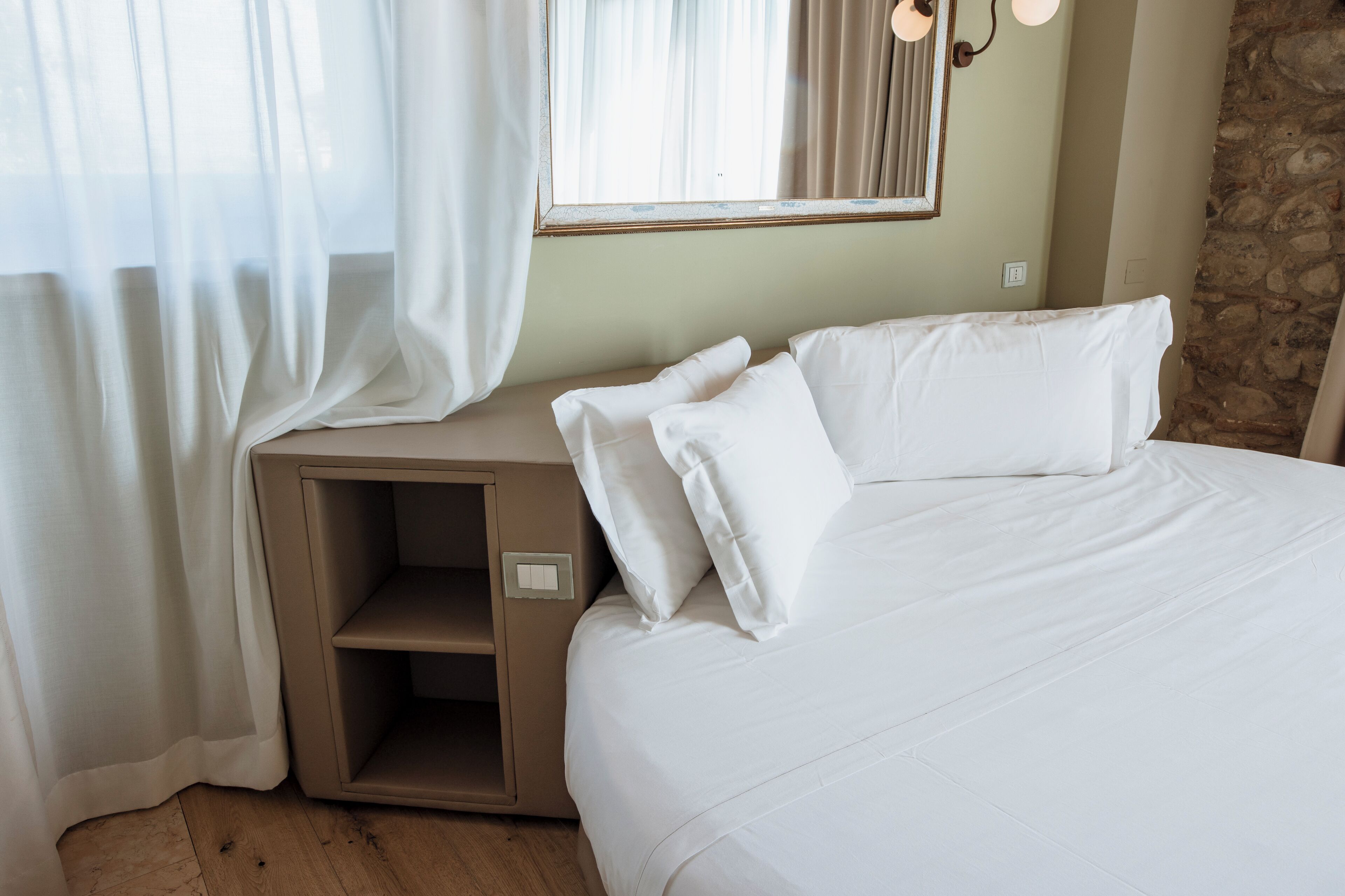 Un lit bien fait, avec du linge de lit blanc, une table de chevet avec une lampe et un mur en pierre.