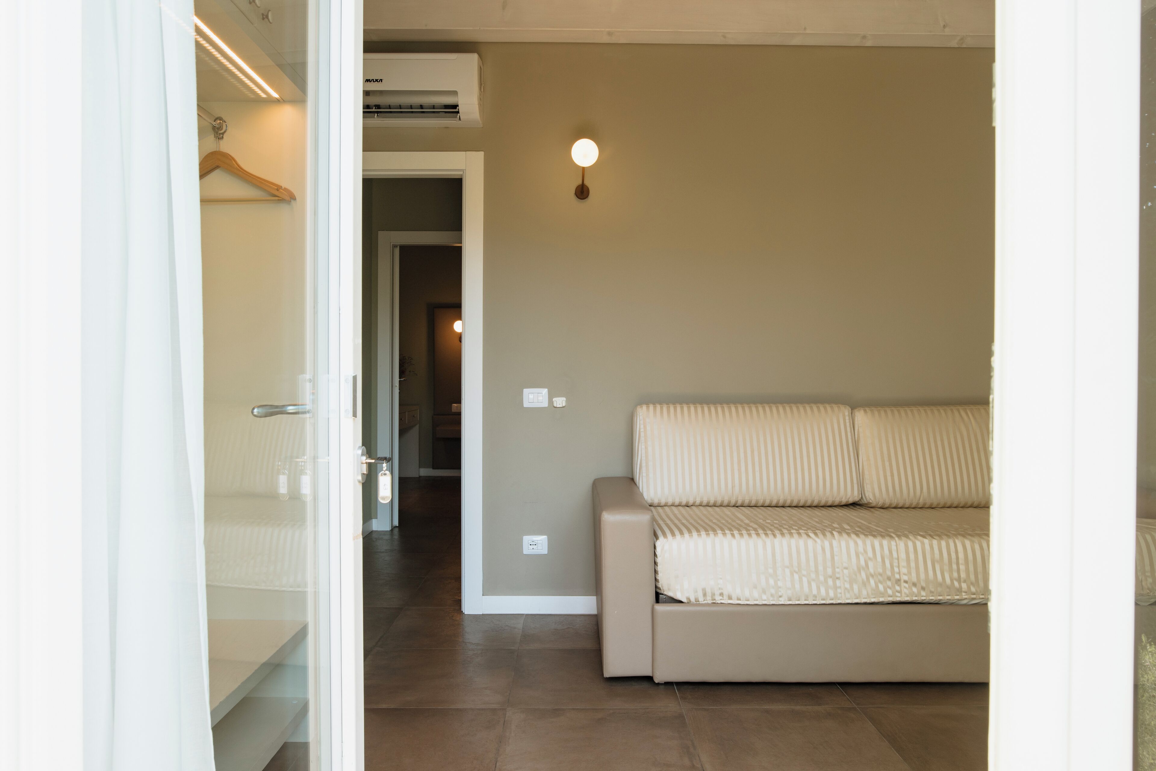Une pièce avec un canapé beige, une porte en verre donnant sur un couloir et un luminaire fixé au mur.