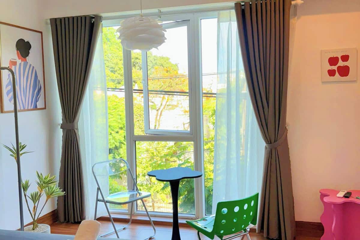 A hotel room with a bed, a small table, a chair, a potted plant, and a view of greenery outside.