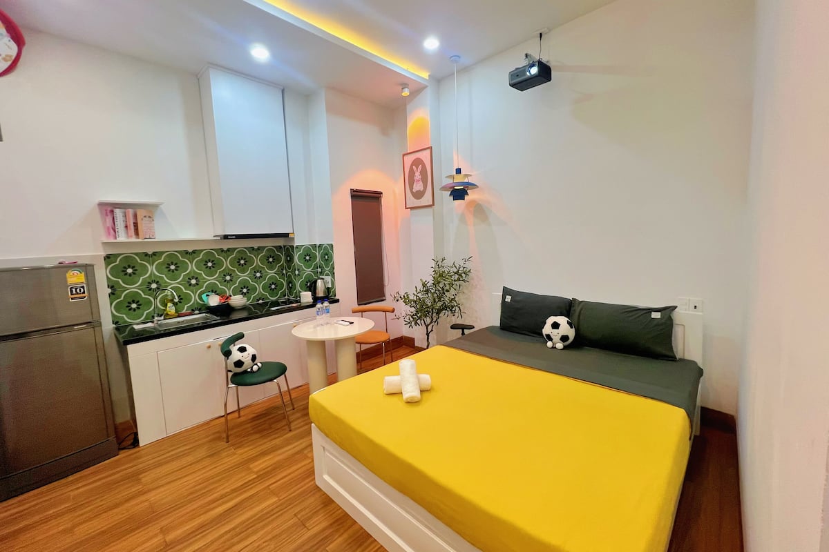 A compact room with a bed, a small kitchenette, a dining area, and a projector.