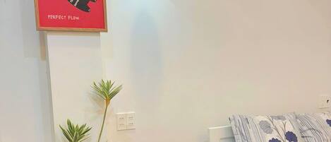 A small room with a bed, a red chair, a white table with a remote control, a hanging light, and a framed poster on the wall.