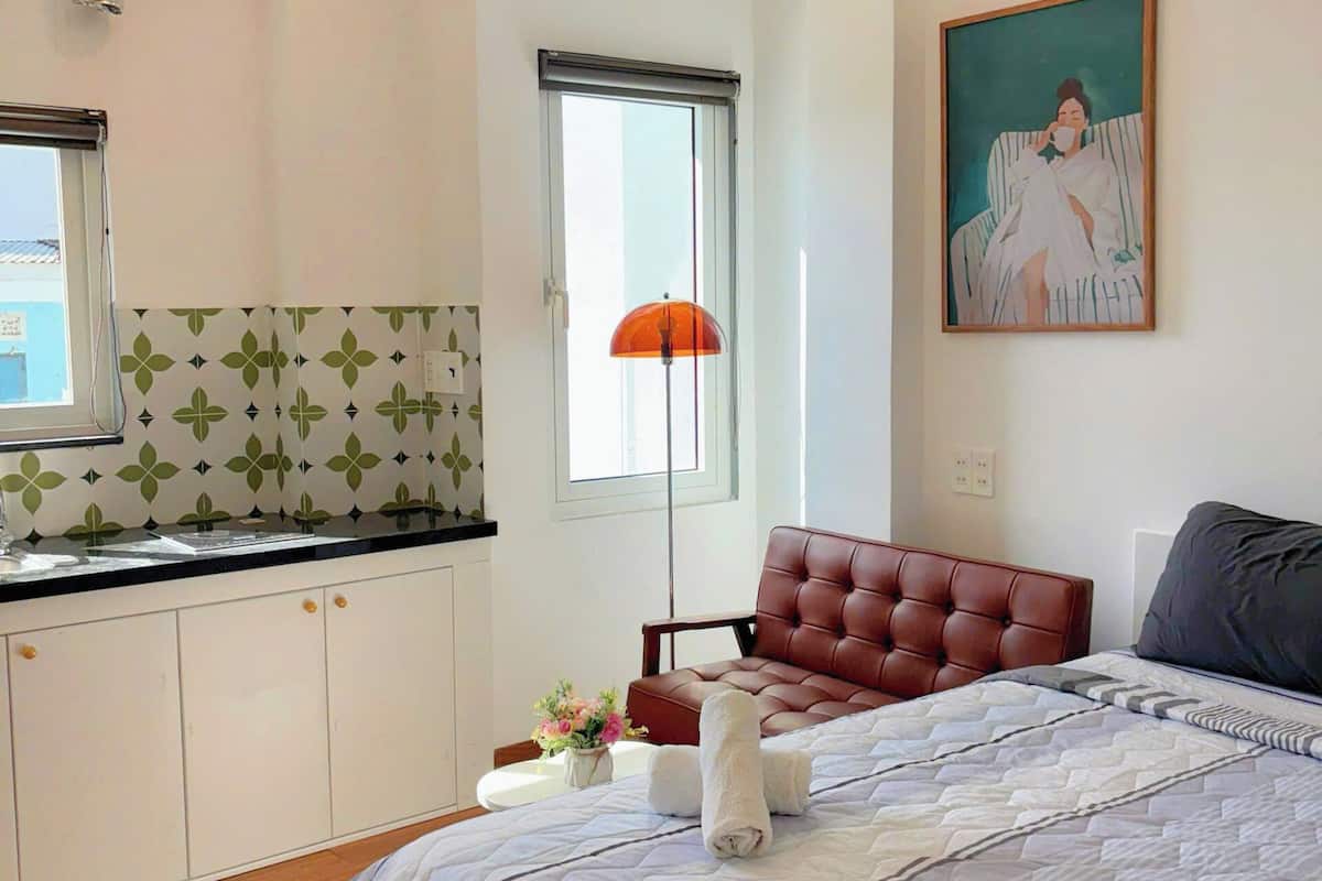 A modern bedroom with a large bed, a separate seating area, a kitchen with a window, and a painting on the wall.