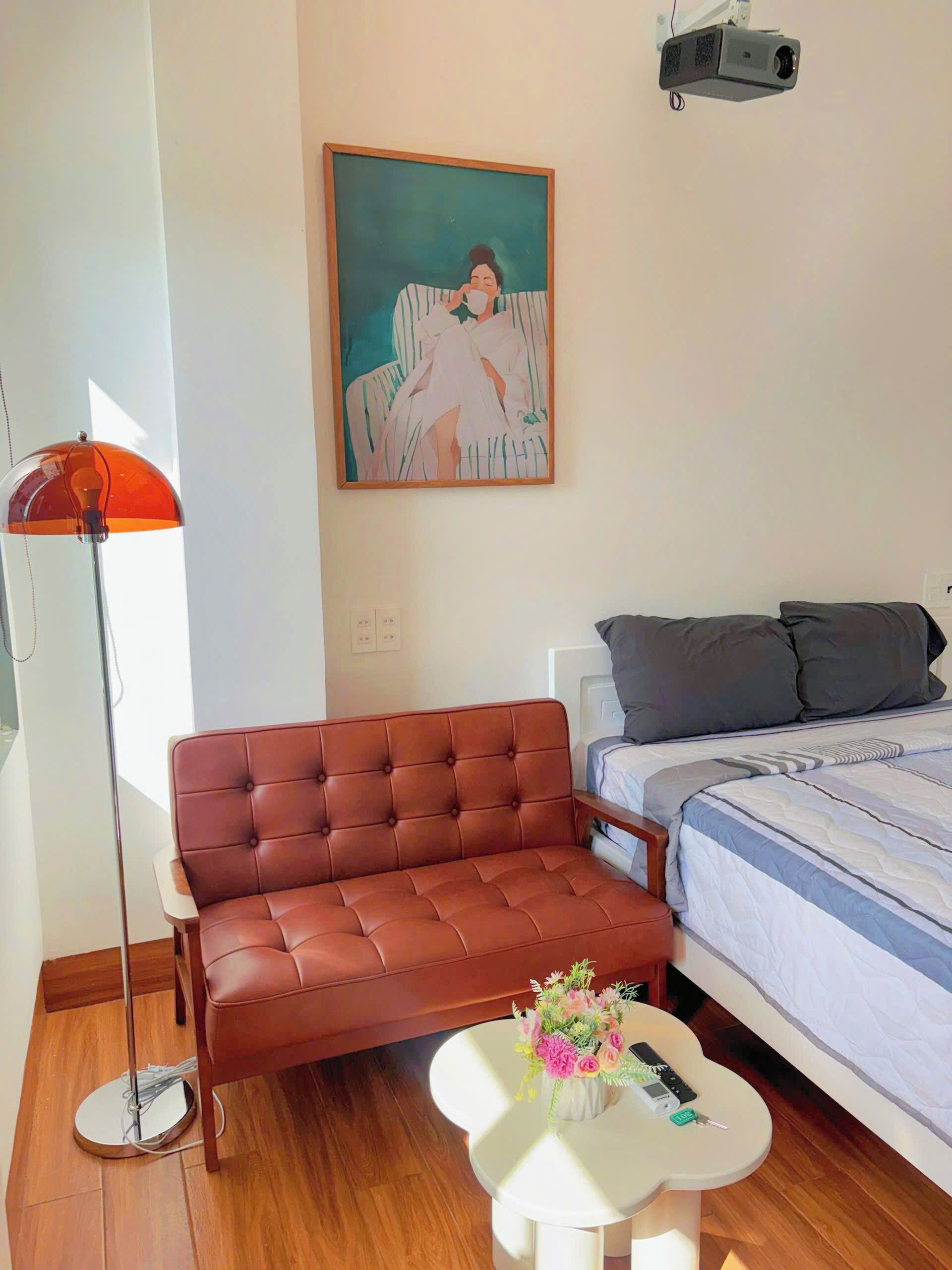 A modern bedroom with a brown leather sofa, a white bedside table with a vase of flowers, a bed with grey bedding, and a painting on the wall.