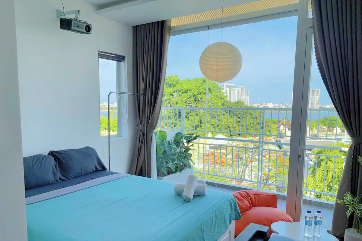 A hotel room with a large bed, a small table, a projector, and a balcony with a view of the city and greenery.