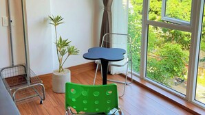 A room with a large window, a potted plant, a green chair, a small table, and a metal chair. - Corgi House - Steps from Dragon Bridge (Da Nang)