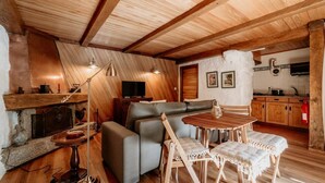 A cozy cabin interior with a wooden ceiling, a fireplace, a sofa, a dining table with chairs, and a kitchen area. - Alojamento T1 Gerês-Junto ao Rio (São Miguel)
