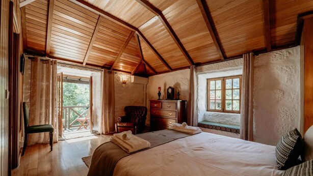 A cozy room with a wooden ceiling, a large bed, a chair, a dresser, and a window with a view of greenery. - Alojamento T1 Gerês-Junto ao Rio (São Miguel)