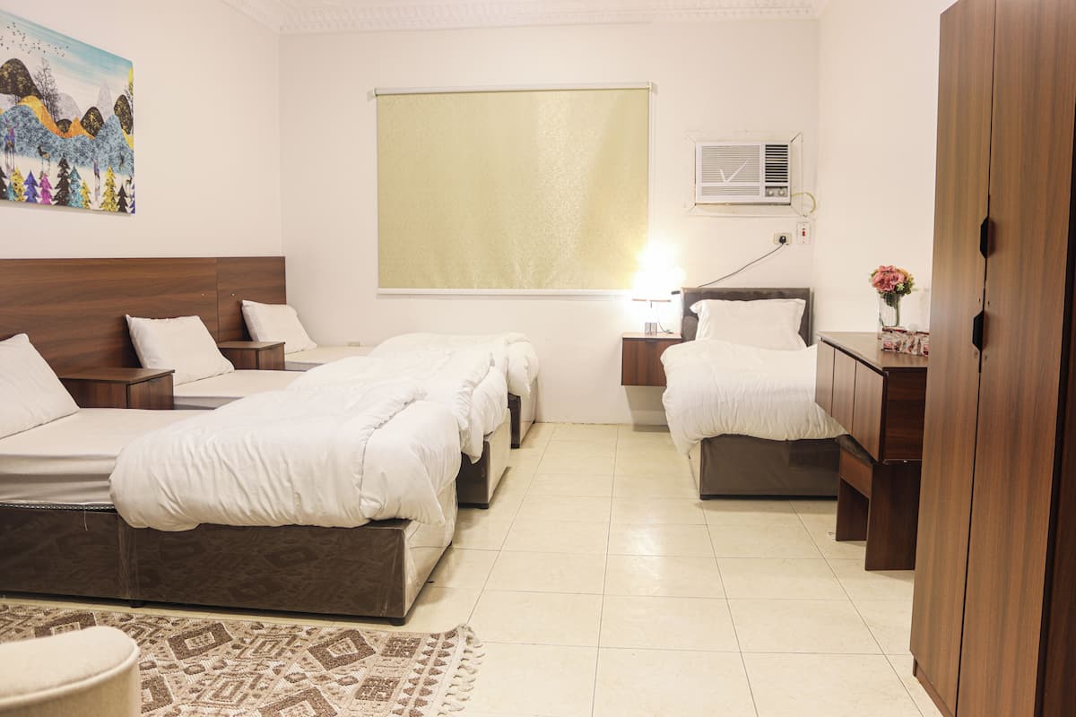 A hotel room with two beds, a wooden wardrobe, a bedside table with a lamp, a window with a blind, and a painting on the wall.