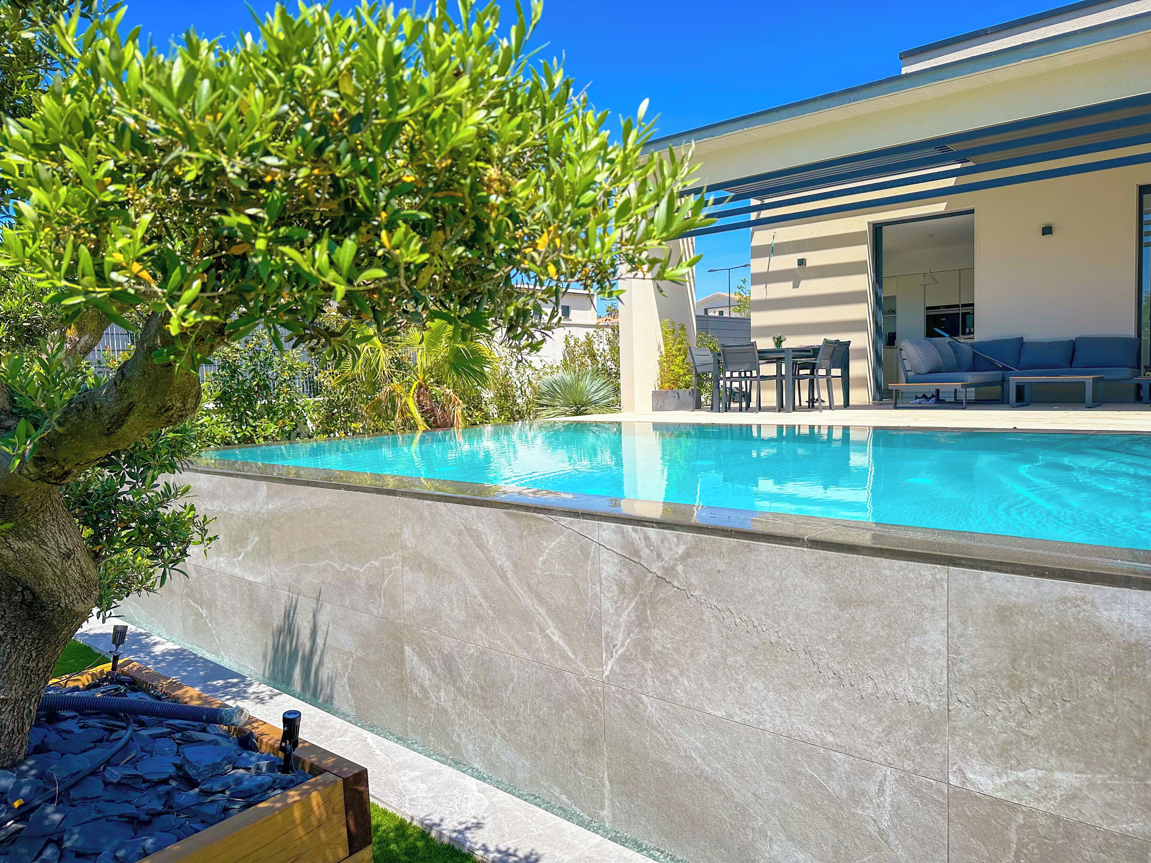 Una piscina con borde de piedra, rodeada de vegetación y un edificio moderno con terraza.