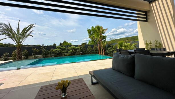 Et moderne poolområde udendørs med en sofa og et bord, omgivet af grøn vegetation og en klar himmel.