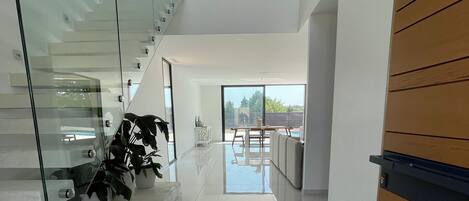 A modern interior with a glass staircase, a wooden door, and a view to an outdoor area.