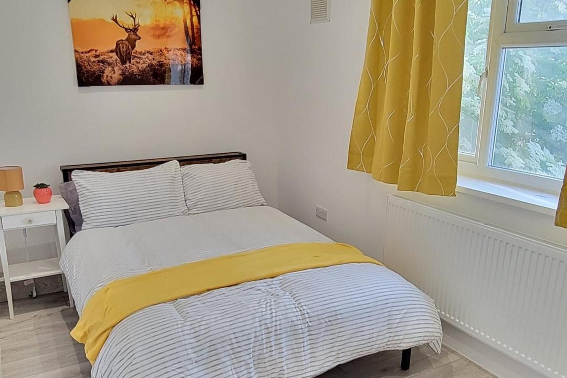 Un dormitorio con cama, mesita de noche, lámpara, ventana con cortinas e imagen colgada en la pared.