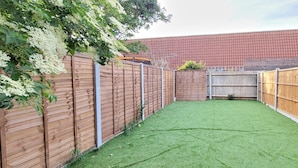 A fenced backyard with artificial grass, a wooden fence, and a brick building in the background.