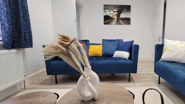 A modern living room with two blue sofas, one with a yellow pillow and the other with a white pillow. A white vase with dried plants is on the floor.
