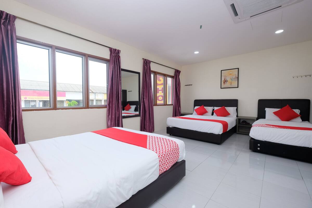 A hotel room with three beds, each with white and red bedding, and large windows with purple curtains.