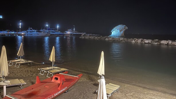 A nighttime marina with boats docked, a rocky breakwater, and beach chairs and umbrellas on the sand.