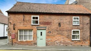 Un bâtiment en briques avec une porte verte, deux fenêtres grillagées et un toit recouvert de tuiles.