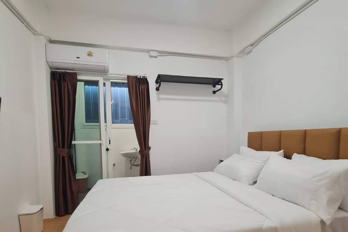 Un dormitorio con cama, una ventana con cortinas, una mesita de noche y un estante fijado a la pared.