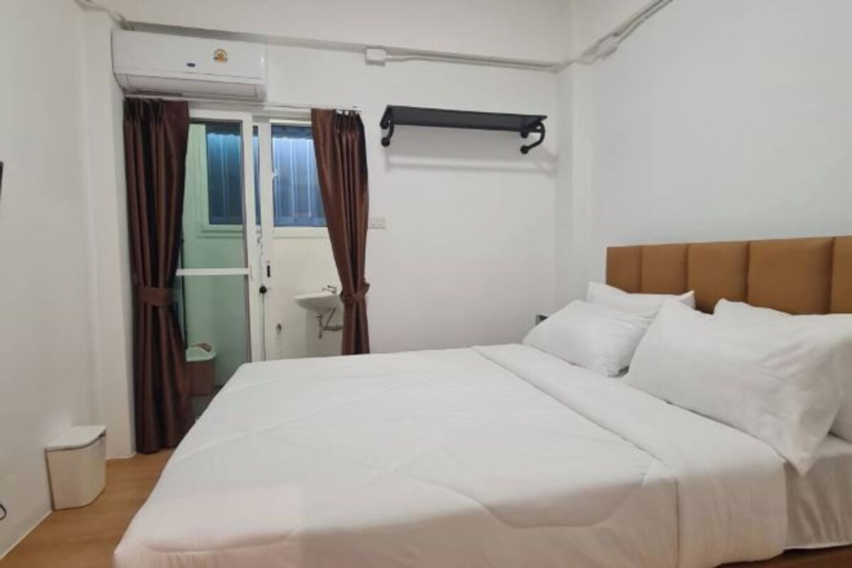 Ein Hotelzimmer mit Bett, einem Fenster mit Vorhängen, einem Nachttisch und einem an der Wand montierten Regal.
