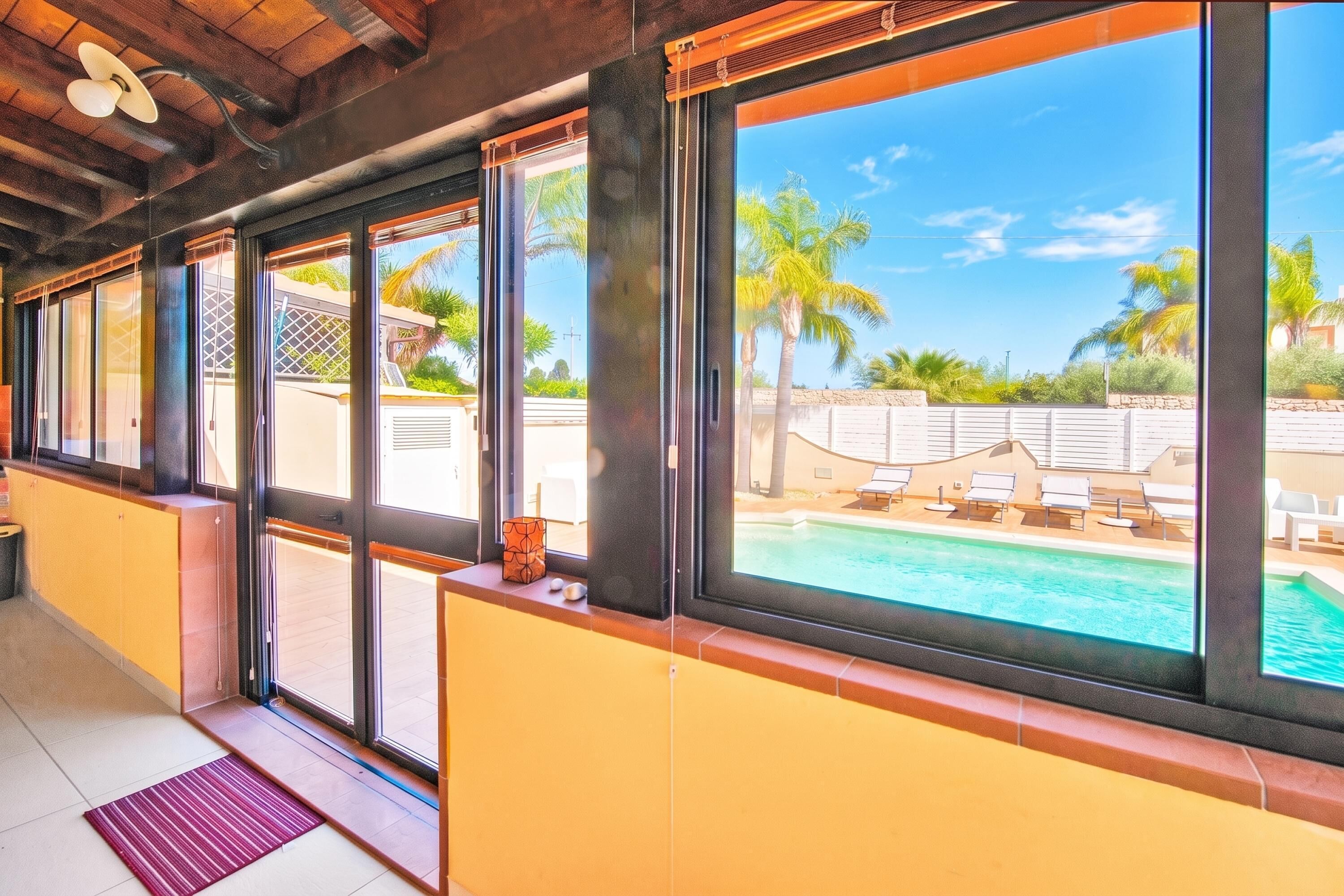 A hotel room with large windows overlooking a pool area with lounge chairs and palm trees.