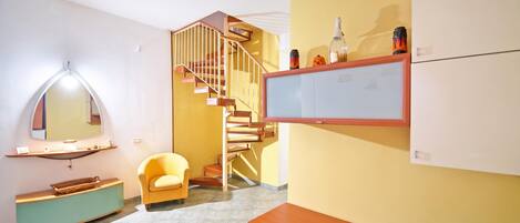 A room with a wooden staircase, a yellow wall, a wooden bench, a chair, and a flat-screen TV mounted on the wall.